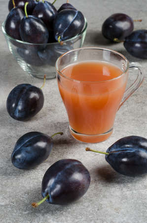 Plum juice in a glass mug and ripe plums in a plate on a gray table.の写真素材