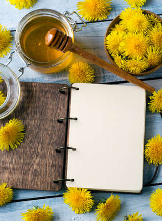 Dandelion honey in jar with fresh yellow flowers on light blue wooden background and notebook with blank page for your inscriptions. Top view.の写真素材