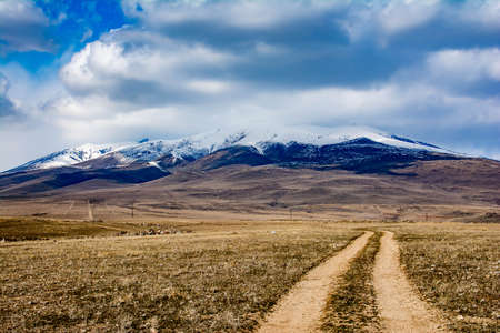 A dirt road leads up the mountain. White clouds over the mountain. The top of the mountain is...