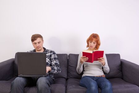 A young couple is sitting on the sofa. She is reading on he is working on his laptop.の写真素材