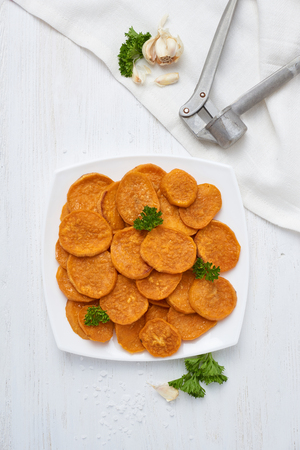 Baked sweet potatoes in white plate on a white wooden table with parsley, garlic and salt. White wooden rustic picnic table scenery from above.の写真素材
