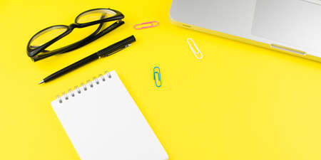 Notepad, pen, black glasses, colorful clips and laptop on yellow background. Flat lay with copy space. Workplace in the office.の写真素材