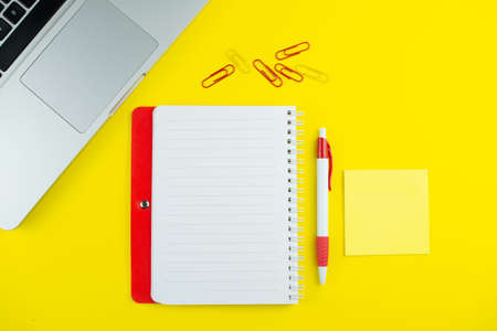 Red notepad, pen, colorful clips and laptop on yellow background. Flat lay with copy space....