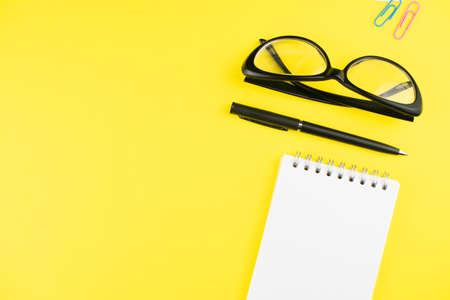 Black glasses, pen, notepad and colorful clips on yellow background. Flat lay. Copy space. Workplace...