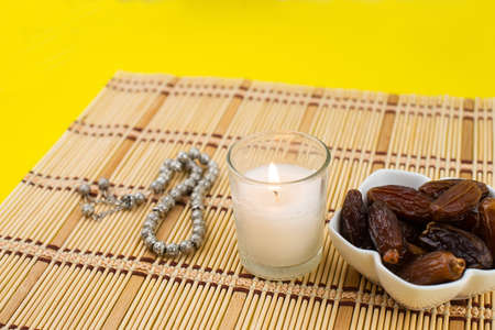 Ramadan Kareem Festival. Close up of dried dates fruit in bowl with candle and rosary on yellow background. Copy spaceの写真素材
