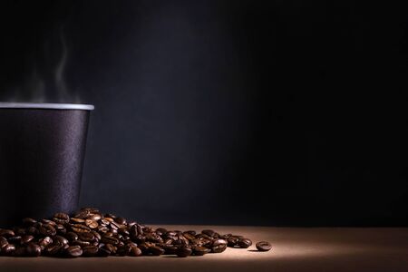 Black disposable cup of coffee with smoke and scattered coffee beans on dark background. Space for textの写真素材