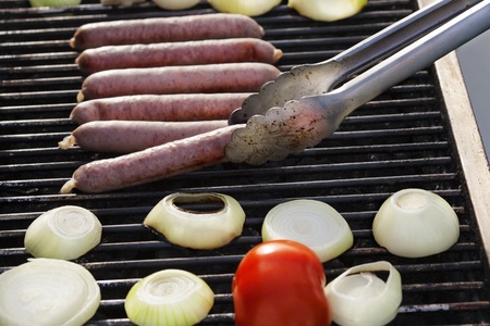 Sausages, onion slices and tomato getting ready on an outdoor barbecue grill. One of the sausages is apparently ready, as tongs are lifting it off of the grill.の写真素材