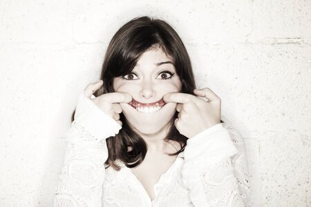 Portrait of a beautiful young woman making a funny face; herfingers pulling her mouth as upwards as possible.の写真素材