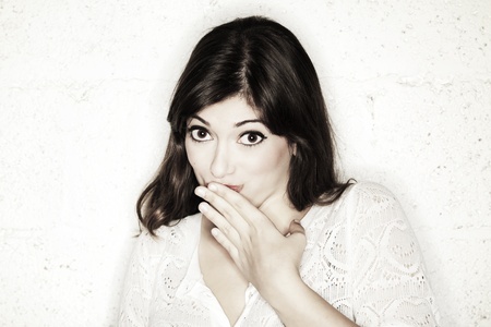Portrait of a beautiful young woman looking at the camera with her hand covering her mouth. Her posture and facial expression suggest she is feeling shy towards something, or maybe guilty. Either way, she is very cute.の写真素材