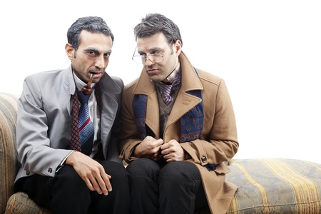 Two adult man (mid 30's and mid 20's) wearing old-man clothes and makeup, sitting on a used up vintage sofa. One of them is looking at the camera with a pipe in his mouth while the other one is looking at him through his vintage glasses. Isolated on whiteの写真素材