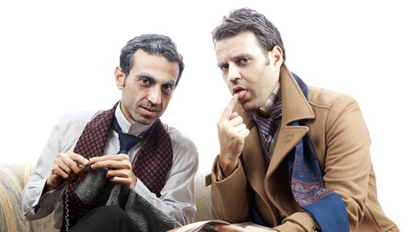Two adult man (mid 30's and mid 20's) wearing old-man clothes and makeup, sitting on a used up vintage sofa. One of them is knitting while the other onewetting his index finger with his tongue in order to flip the page of the newspaper he's holding. Isolaの写真素材