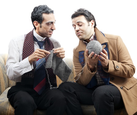 Two adult man (mid 30's and mid 20's) wearing old-man clothes and makeup, sitting on a used up vintage sofa. One of them is knitting while the other one is holding the ball of yarn. They appear to be in the middle of a conversation. Isolated on white backの写真素材
