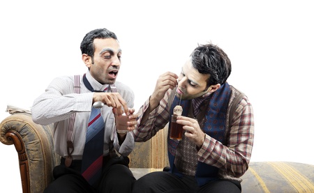 Two adult man (mid 30's and mid 20's) wearing old-man clothes and makeup, sitting on a used up vintage sofa. The left one is very anxious to get the teabag, so he's concentrated on the right one's actions taking out the teabag out of the glass in an awkwaの写真素材