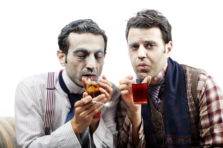 Two adult man (mid 30's and mid 20's) wearing old-man clothes and makeup, sitting on a used up vintage sofa. Looking rather cold and pitiful they are both warming themselves with a glass of hot tea. Isolated on white background.の写真素材