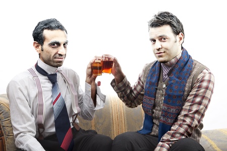 Two adult man (mid 30's and mid 20's) wearing old-man clothes and makeup, sitting on a used up vintage sofa. Both of them are smiling at the camera while toasting their tea glasses. Isolated on white background.の写真素材