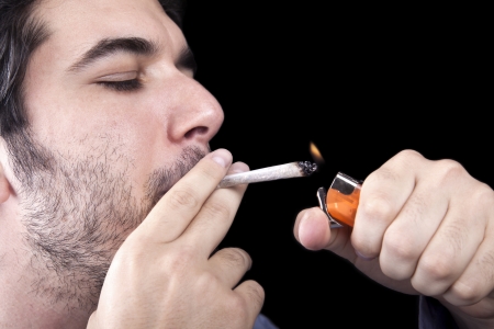 Closeup of an adult man (30 years old) with his profile to the camera. He appears to be quite a bum, being unshaved and wearing a light blue fabric robe, concentrated in lighting a marijuana jspliff (aka reefer; joint) with a simple disposable half-transpの写真素材
