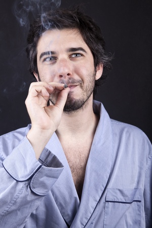 Medium closeup of an adult man (30 years old), which  appears to be quite a bum, being unshaved and wearing a light blue fabric robe, looking upwith a satisfied look while smoking a marijuana spliff (aka reefer; joint). Dark gray background.の写真素材