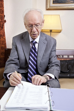 An elderly (in his 80's) business man wearing suit and tie sitting in a hotel's business lounge, going over some papers with budget details.の写真素材