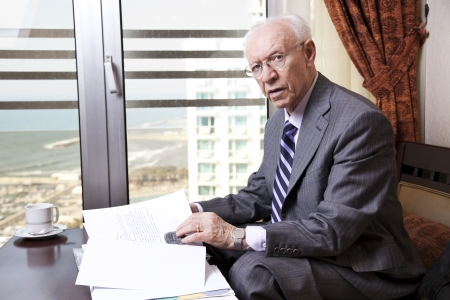 An elderly (in his 80's) business man wearing suit and tie sitting in a hotel's business lounge, looking at camera, as if disturbed in the middle of going over some papers after having coffee. The sea and a marina can be seen defocused in the background.の写真素材