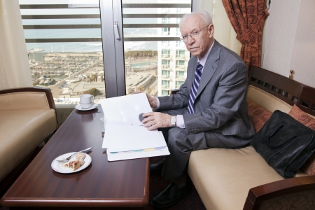 An elderly (in his 80's) business man wearing suit and tie sitting in a hotel's business lounge, looking at camera, as if disturbed in the middle of going over some papers after having coffee. The sea and a marina can be seen defocused in the background.の写真素材