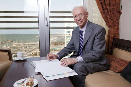 An elderly (in his 80's) business man wearing suit and tie sitting in a hotel's business lounge, looking at camera with a slight smile on his face, in the middle of going over some papers after having coffee and cake. The sea and a marina can be seen defoの写真素材