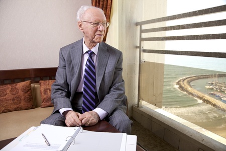 An elderly (in his 80's) business man wearing suit and tie sitting in a hotel's business lounge, looking through the window for a little break from the documents he is inspecting.の写真素材