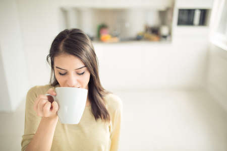 Young woman enjoying,holding cup of hot beverage,coffee or tea in morning sunlight.Enjoying her morning coffee in the kitchen.Savoring a cup of coffee breathing in the aroma in bliss and appreciationの写真素材