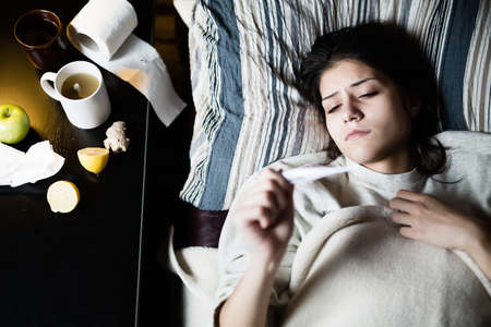 Sick young woman in bed at home having flu,measuring temperature.Thermometer to check her...
