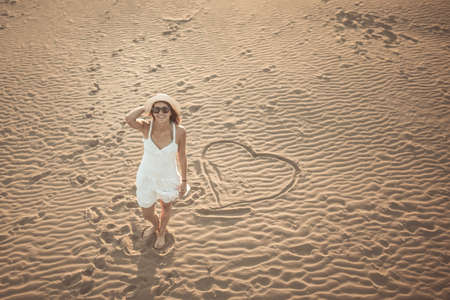 Woman on the beach making heart on the sand.Young woman walking on the sand in a white dress.Relaxed woman breathing fresh air.Travel and vacation.Freedom,inspiration,love.Birds eye view.Valentinesの写真素材
