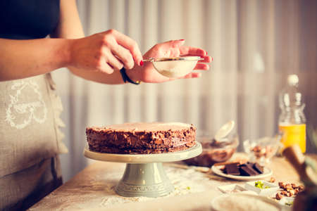 House wife wearing apron making finishing touches on birthday dessert chocolate cake.Woman making homemade cake with easy recipe,sprinkling powdered sugar on top.Icing sugar sprinkled with colanderの写真素材