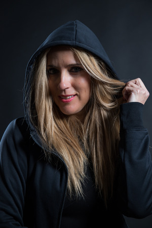 Woman with blonde hair and blue eyes dressed in dark color and with her head covered, she poses in a photographic studio with black background.の写真素材