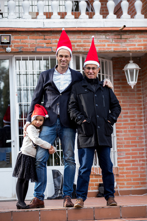 Grandfather, father and daughter wearing a Santa cap in front of their house