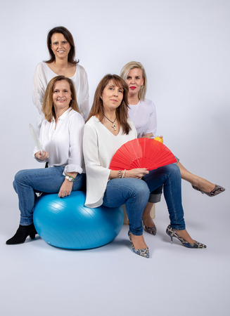 Group of 4 women, friends, middle-aged having fun in a photo session in a studio with white background, sitting on a ball and playing with a fanの写真素材