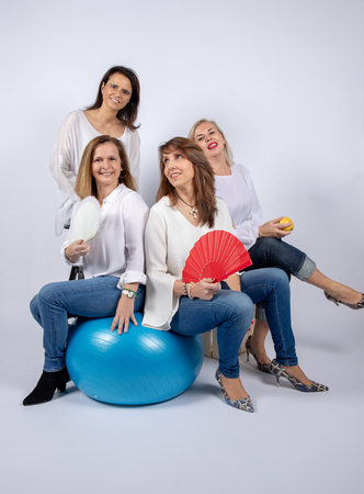 Group of 4 women, friends, middle-aged having fun in a photo session in a studio with white background, sitting on a ball and playing with a fanの写真素材