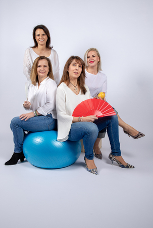 Group of 4 women, friends, middle-aged having fun in a photo session in a studio with white background, sitting on a ball and playing with a fanの写真素材