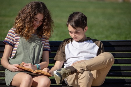 A couple of siblings quietly read a book sitting on a bench on the promenade.
