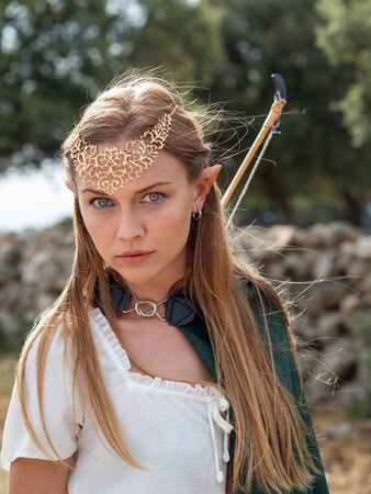 Blonde girl with blue eyes and makeup with elf ears poses in the field with a bow and a green cape.の写真素材