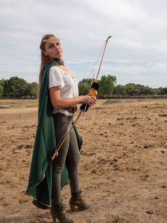 Blonde girl with blue eyes and makeup with elf ears poses in the field with a bow and a green cape.の写真素材