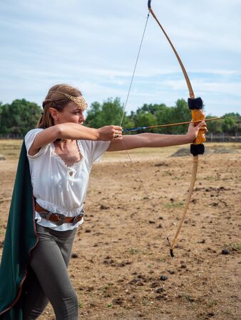 Blonde girl with blue eyes and makeup with elf ears poses in the field with a bow and a green cape.の写真素材
