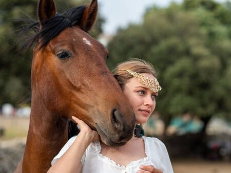 Blonde girl with blue eyes and makeup with elf ears and a tiara on her head poses in the field with a brown horse and a green cape.の写真素材
