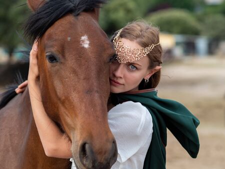 Blonde girl with blue eyes and makeup with elf ears and a tiara on her head poses in the field with a brown horse and a green cape.の写真素材