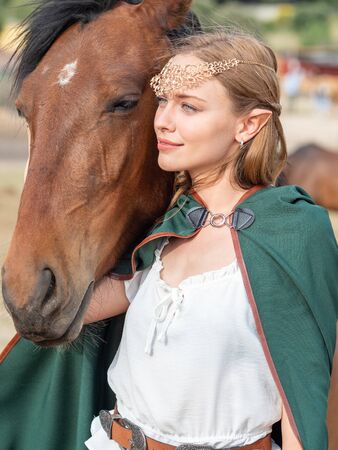Blonde girl with blue eyes and makeup with elf ears and a tiara on her head poses in the field with a brown horse and a green cape.の写真素材