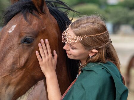 Blonde girl with blue eyes and makeup with elf ears and a tiara on her head poses in the field with a brown horse and a green cape.の写真素材