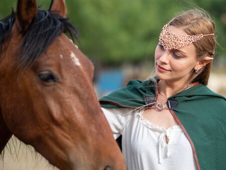 Blonde girl with blue eyes and makeup with elf ears and a tiara on her head poses in the field with a brown horse and a green cape.の写真素材