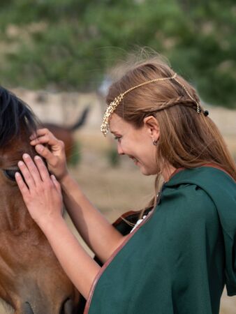 Blonde girl with blue eyes and makeup with elf ears and a tiara on her head poses in the field with a brown horse and a green cape.の写真素材