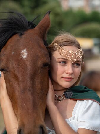 Blonde girl with blue eyes and makeup with elf ears and a tiara on her head poses in the field with a brown horse and a green cape.の写真素材