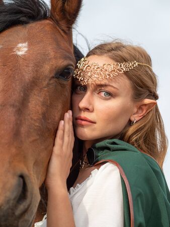 Blonde girl with blue eyes and makeup with elf ears and a tiara on her head poses in the field with a brown horse and a green cape.の写真素材