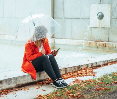 Caucasian girl with red gabardine walks through a park under a transparent umbrella.の写真素材