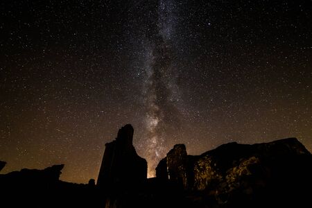Long exposure nocturnal photograph with the Milky Way in the background, between mountains and next to a building with illuminated windows.の写真素材