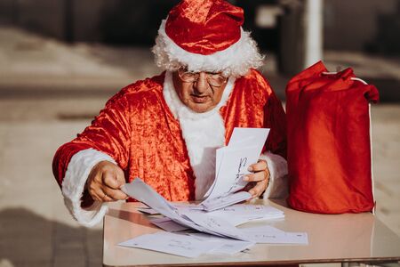 Stock photo of Santa claus without beard sitting on a terrace organizing Christmas cards with a red bag on the table. Christmas timeの写真素材